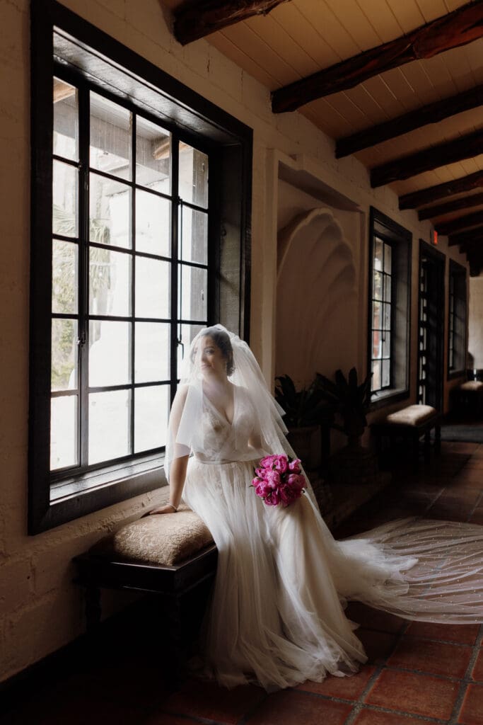 A bride sits on a bench by a large window in a dimly lit hallway. She wears a flowing white gown and veil, holding a bouquet of pink flowers. Natural light softly illuminates her and the rustic interior, captured perfectly by a San Antonio wedding photographer.