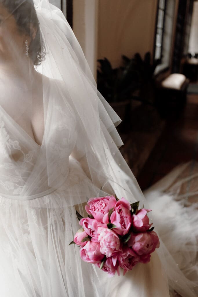 A bride in a white wedding gown and veil holds a bouquet of vibrant pink peonies. The background features soft lighting and elegant architecture, creating a romantic atmosphere, beautifully captured by a talented McAllen wedding photographer.