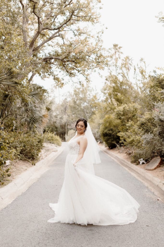 A bride in a flowing white wedding dress stands on a tree-lined path, smiling. She is holding part of her dress in one hand, with a veil draped down her back. Captured by a talented Edinburg wedding photographer, the scene is serene with lush greenery enveloping the path.