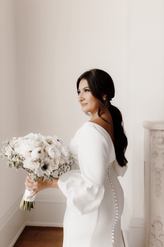 A bride in a white dress stands beside a cream-colored wall, holding a bouquet of white and pink flowers. Her dress features button details along the back, and she has long dark hair styled in loose waves, captured perfectly by a San Antonio wedding photographer.