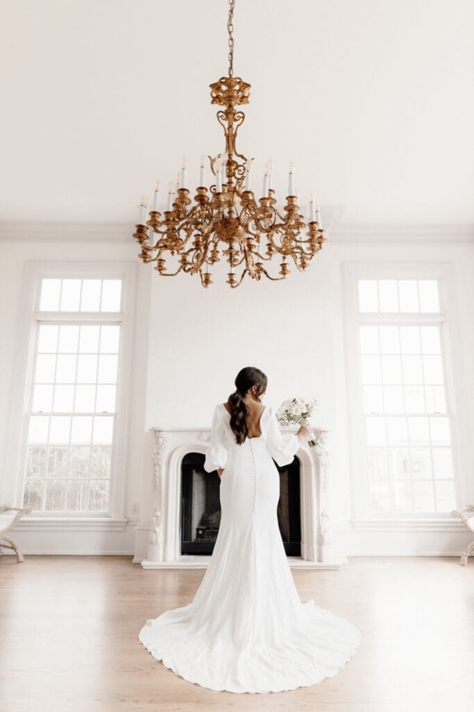 A bride in a flowing white dress stands in an elegant room with a large, ornate chandelier, captured by an Edinburgh wedding photographer. She faces a white fireplace and holds a bouquet of flowers. Two large windows frame the scene, adding natural light to the space.