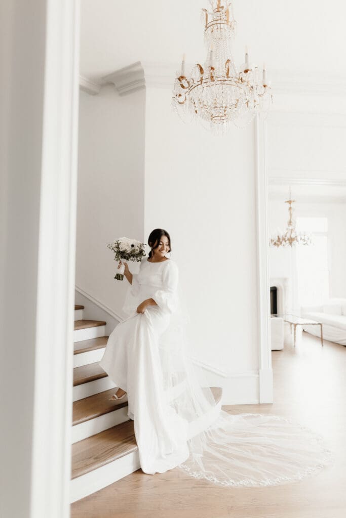 A bride in a white gown holding a bouquet descends a wooden staircase. The room, captured by a McAllen wedding photographer, is elegant with a large chandelier and minimalist decor. Light streams in, highlighting her dress and veil.