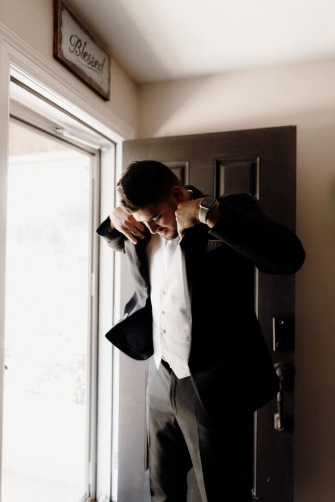 A person dressed in a suit adjusts their jacket near a doorway, with sunlight streaming in. A small sign reading Blessed hangs above the door. The interior is softly lit, creating a warm atmosphere, perfect for capturing by a wedding photographer in McAllen.