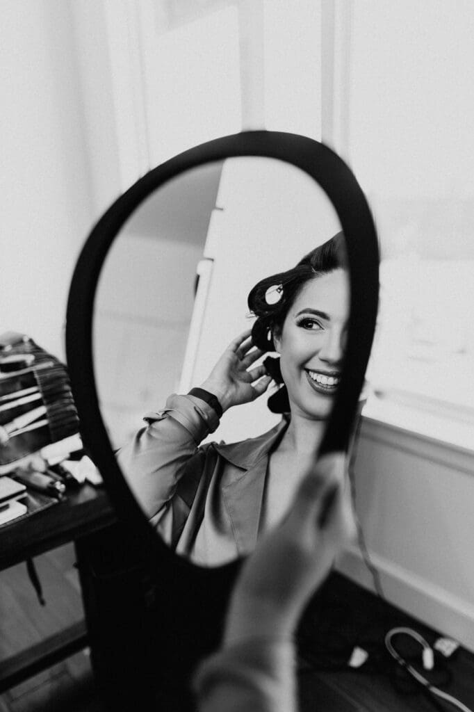 A woman with hair rollers smiles at her reflection in a hand-held mirror. Captured in black and white by a skilled San Antonio wedding photographer, the scene reveals a table of styling tools beside her against a bright window.