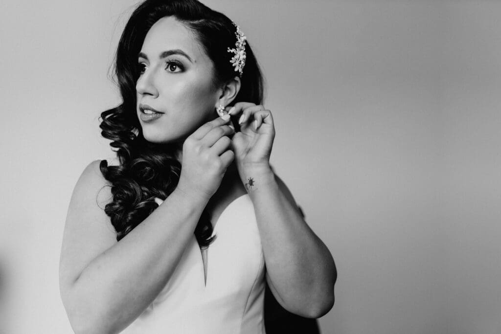 Black and white photo of a woman in a wedding dress, adjusting an earring. Captured by a San Antonio wedding photographer, she has long, wavy hair with an ornate hairpiece and gazes thoughtfully to the side against a plain background.