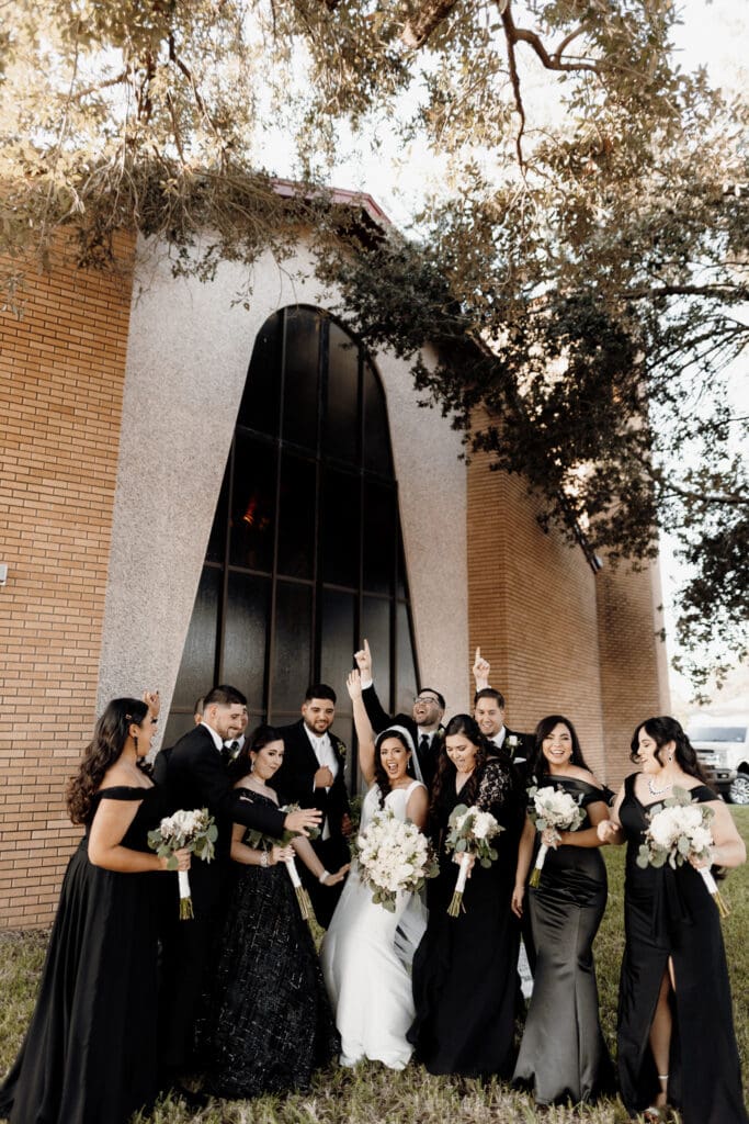 A bride and groom stand with a wedding party of ten, elegantly dressed in formal black attire, in front of a building with a large arched window. Captured by a talented Edinburg wedding photographer, the group smiles brightly with bouquets in hand, framed by trees above.