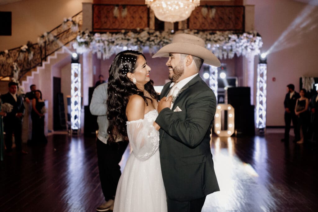 A bride and groom smile at each other while dancing, captured perfectly by a South Padre Island wedding photographer. The bride wears a white, off-shoulder gown, and the groom sports a dark suit with a cowboy hat. The room is elegantly decorated with flowers and lights, guests visible in the background.