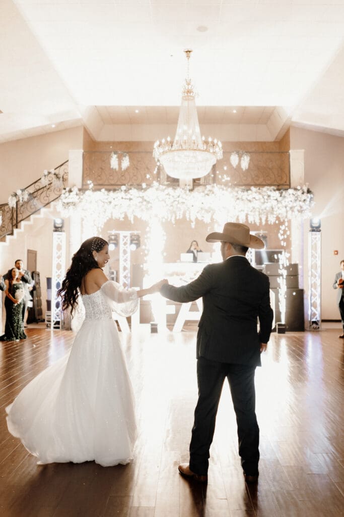 A bride and groom dance under a grand chandelier, beautifully captured by a McAllen wedding photographer. The bride is in a white gown, and the groom, in a suit with a cowboy hat, moves gracefully. Elegant floral decorations and stairs frame the scene as guests watch their enchanting dance.