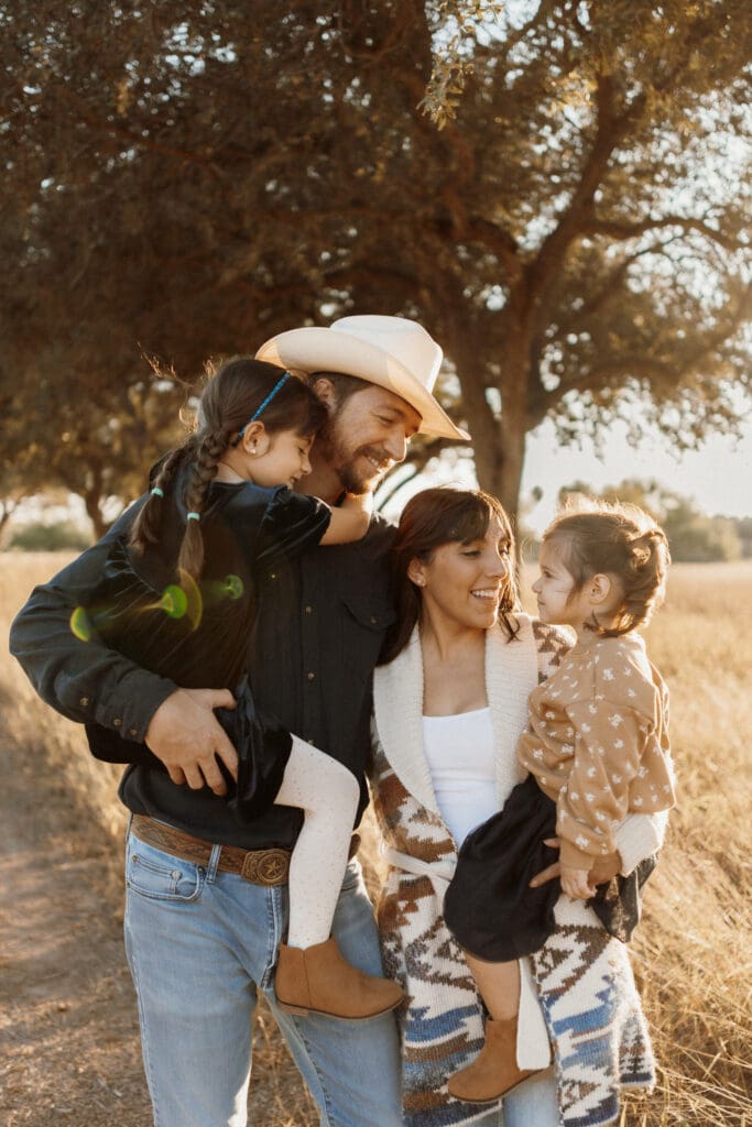 A family of four stands outdoors by a dirt path, surrounded by trees. The man, wearing a cowboy hat, holds a young girl. The woman in a patterned cardigan holds another young girl. Captured by an Edinburg wedding photographer, everyone is smiling and enjoying the sunny day.