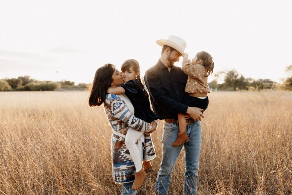 A family stands in a field at sunset, captured beautifully by an Edinburg wedding photographer. A woman holds a child, both in patterned clothing, while a man in a hat embraces another child in polka dots. The scene radiates warmth and serenity, set against the grassy landscape.
