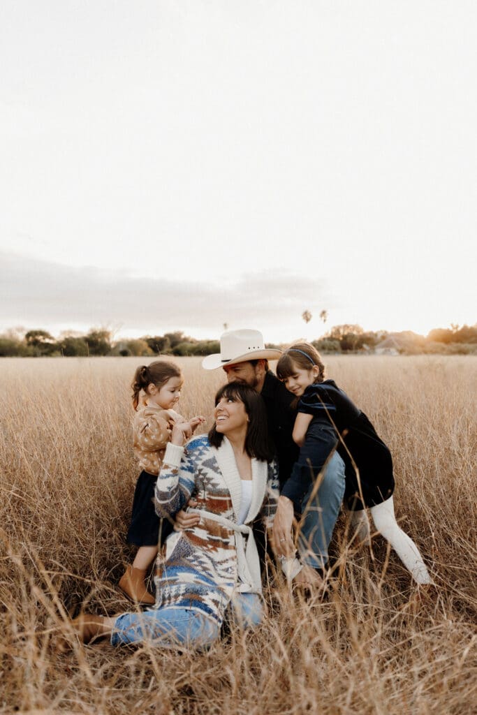 A family sits together in a field of tall grass at sunset, captured beautifully by a McAllen wedding photographer. An adult wearing a patterned sweater and hat kneels, smiling and interacting with two children. The sky is overcast with hints of sunlight in the distance.