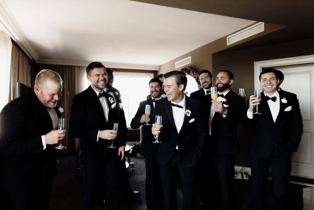 A group of men in tuxedos are sharing a laugh while holding champagne glasses in a warmly lit room. With boutonnières signaling a wedding, they appear relaxed and joyful, making it an ideal scene for a McAllen wedding photographer to capture.