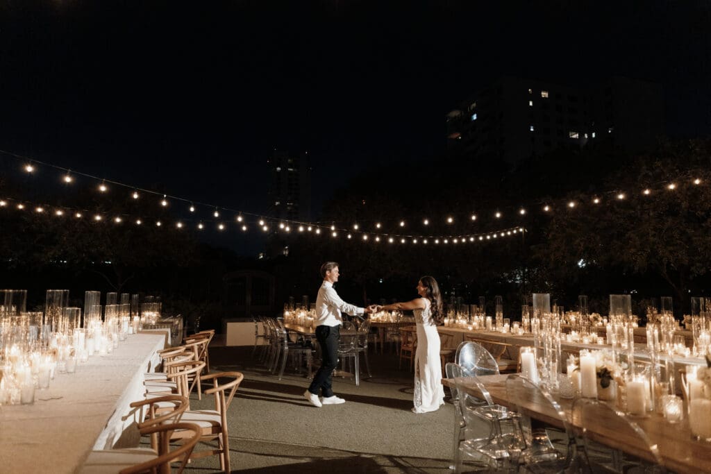 A couple dances under string lights at night, surrounded by tables adorned with numerous lit candles. The atmosphere is romantic and intimate, with trees and tall buildings visible in the background—a perfect scene captured by a talented San Antonio wedding photographer.