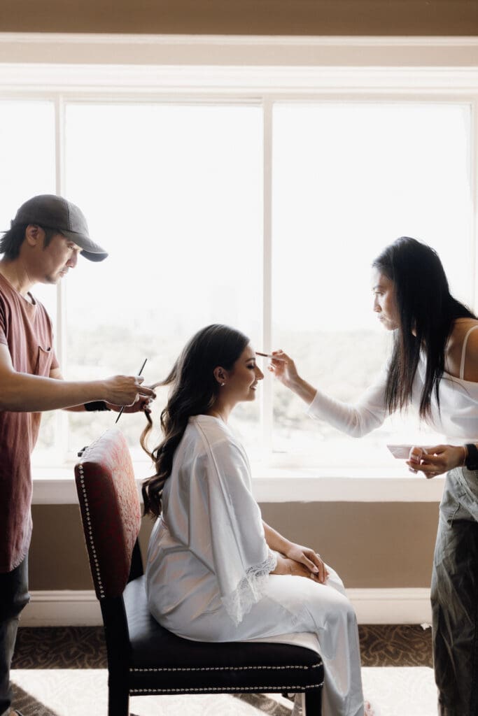 A woman sits in front of a large window while two people style her, preparing for her special day. Her hair is elegantly arranged as makeup is carefully applied. Draped in a light-colored robe on a red chair, she gazes at the blurred greenery outside, captured perfectly by a San Antonio wedding photographer.