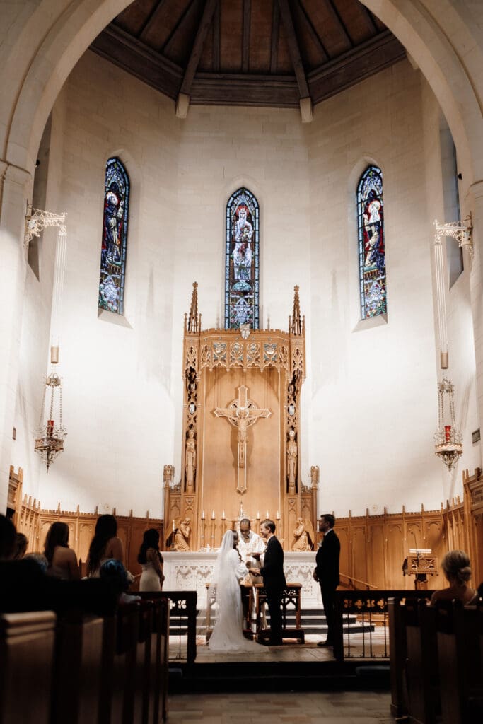 A couple stands at the altar in a grand church with high ceilings, exchanging vows. The backdrop features ornate wooden carvings and stained glass windows, captured beautifully by a San Antonio wedding photographer. Guests are seated on either side, witnessing the ceremony.