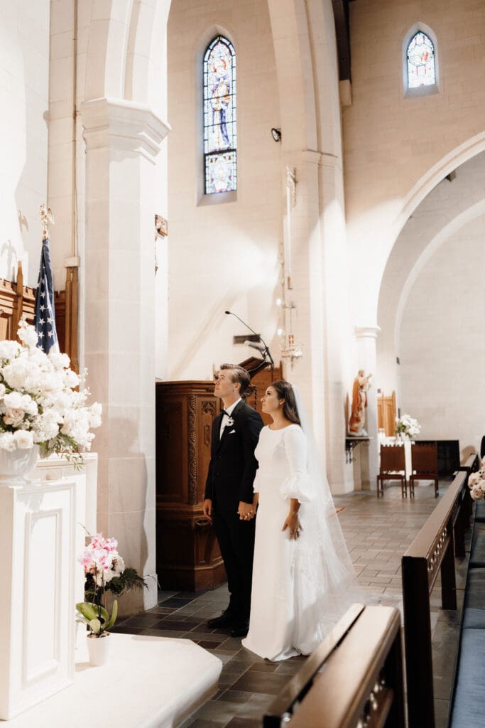 A couple stands at the altar in a church adorned with white flowers and American flags, captured perfectly by a talented San Antonio wedding photographer. The bride wears a white dress and veil, while the groom is in a dark suit. Stained glass windows and arched ceilings are visible in the background.