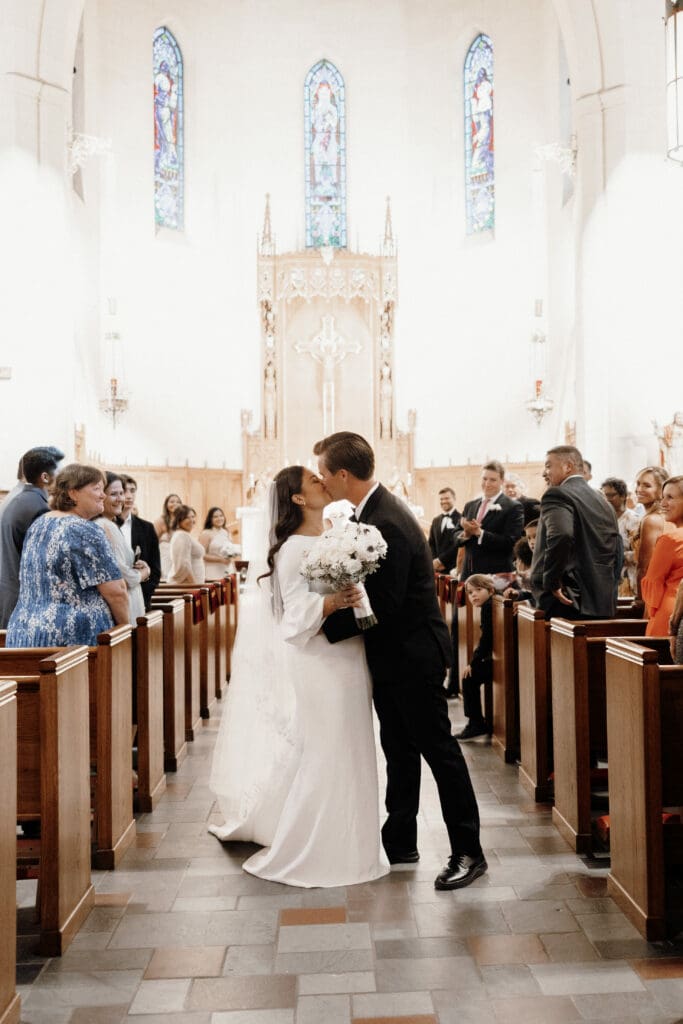 A bride and groom kiss in the aisle of a church after their wedding ceremony, captured beautifully by a McAllen wedding photographer. The bride holds a bouquet of white flowers, and guests stand on either side watching. The church interior is decorated with stained glass windows and ornate designs.