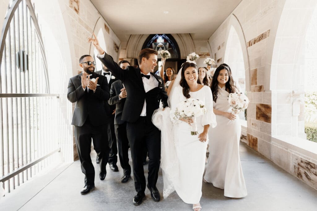 A joyful wedding procession walks down a corridor, captured perfectly by a skilled McAllen wedding photographer. The bride, in a white dress holding a bouquet, smiles alongside the groom in a tuxedo. Behind them, cheering bridesmaids and groomsmen hold flowers, adding to the enchanting moment.