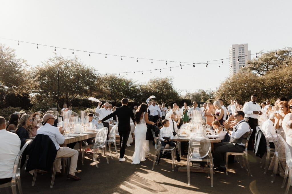 A bride and groom gracefully dance around a long outdoor dining table, their joyful scene captured by a skilled McAllen wedding photographer. Guests applaud under the glow of natural sunlight and string lights. In the background, trees and a tall building complete this idyllic celebration.