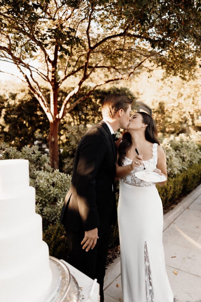 A bride and groom share a kiss outdoors next to a white wedding cake. The bride is in a white dress, and the groom is in a black suit. Captured by an Edinburg wedding photographer, they are surrounded by lush greenery and trees under a sunlit sky.