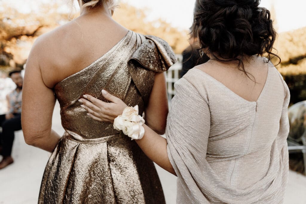 Two women stroll arm in arm outdoors, one in a metallic gold one-shoulder gown and the other in a light beige dress accented by a corsage. Captured as if by an Edinburgh wedding photographer, they appear to be at an elegant formal event.