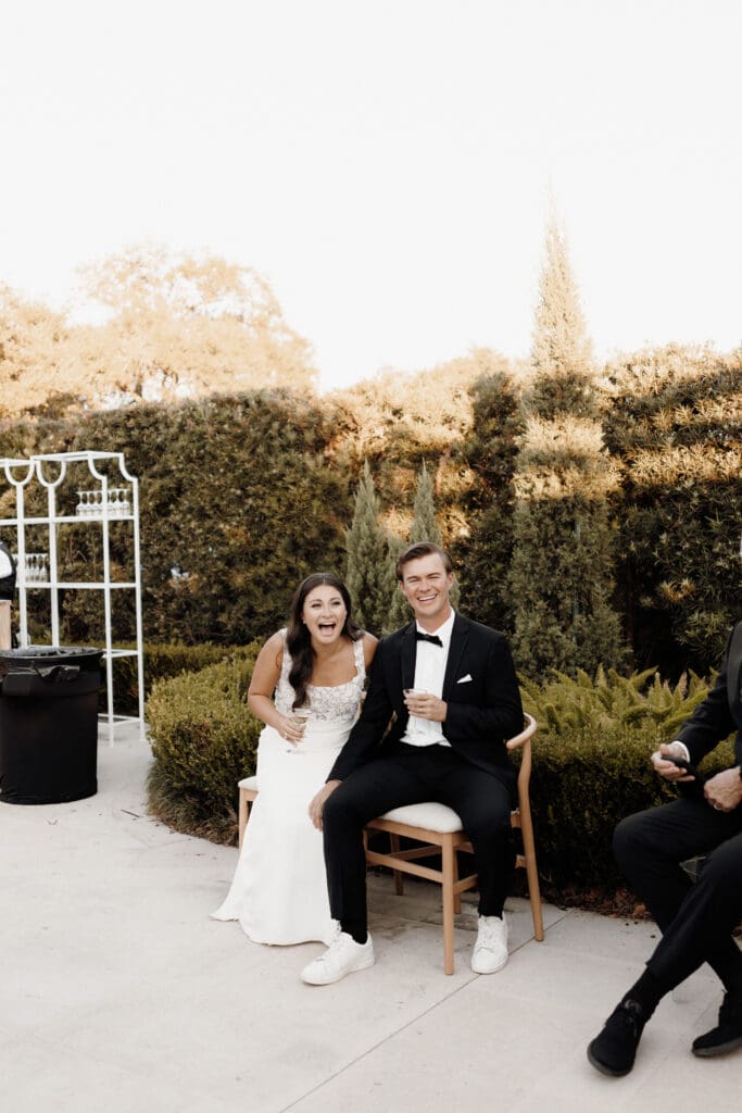 Bride and groom sit outdoors, smiling joyfully. The bride dazzles in a white wedding dress while the groom rocks a black suit with a bow tie and white sneakers. Captured by a talented San Antonio wedding photographer, they are surrounded by lush greenery that adds to the relaxed and festive atmosphere.