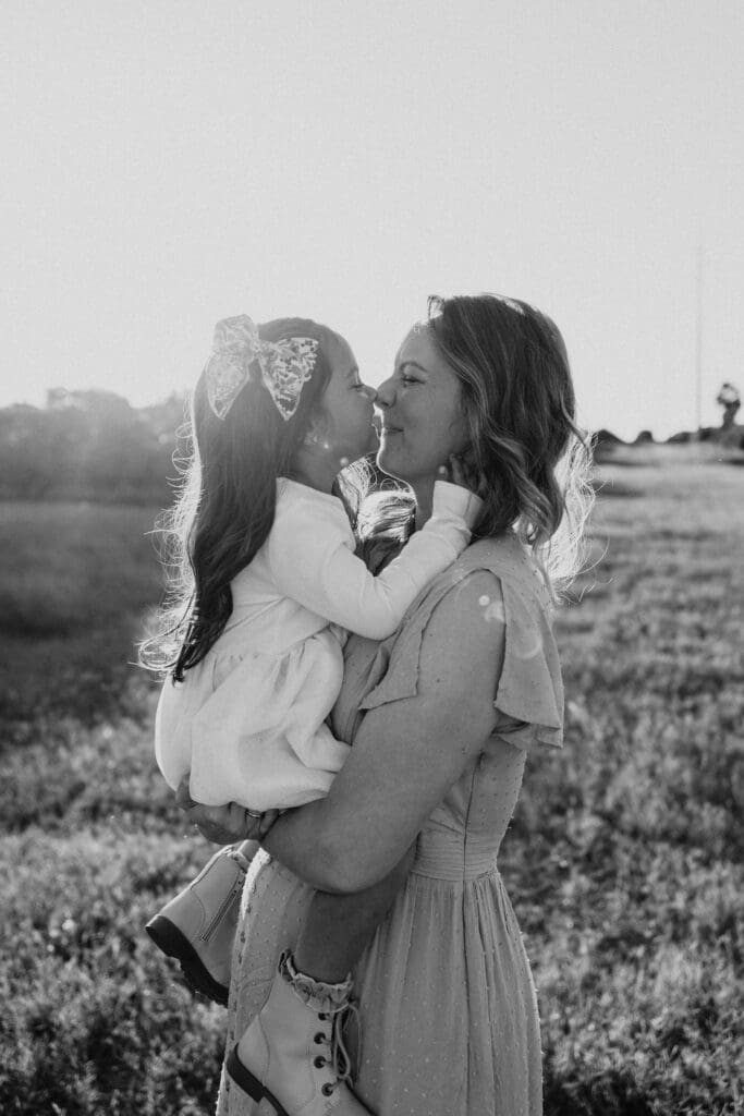 A woman holds a young girl lovingly in a grassy field. The girl, wearing a bow in her hair, leans in for a nose-to-nose touch. Both are dressed in light clothes, and the sunlight creates a warm glow around them—a tender moment captured by an Edinburg wedding photographer. Black and white image.