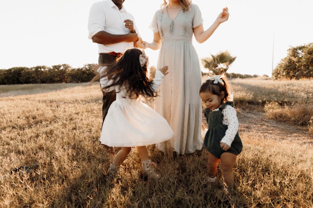 A family stands in a sunny field. A man, woman, and two young girls enjoy a moment together captured by a skilled wedding photographer in McAllen. The girls are wearing dresses and playing on the grass. The background features trees and a bright sky, perfect for a McAllen wedding photographers vibrant shot.