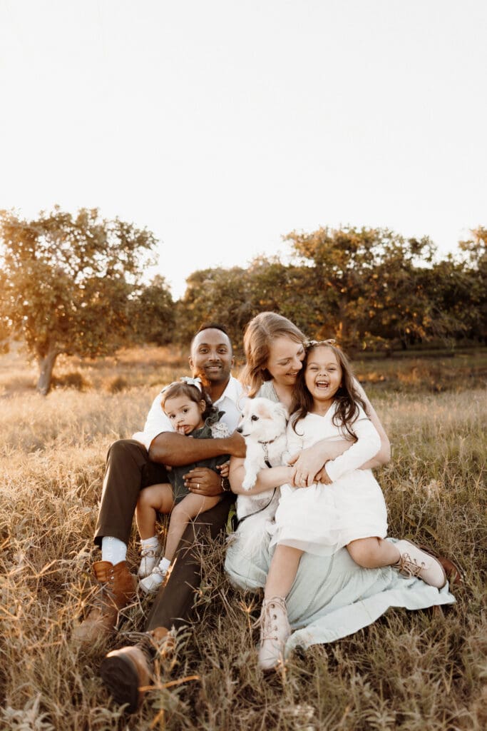 A family of four sits on grass in a sunny field. The parents, each holding a young child, are joined by a white fluffy dog. Everyone is smiling, enjoying the warm, golden hour light. This moment could easily be captured by a skilled wedding photographer in McAllen with trees softly framing the scene.
