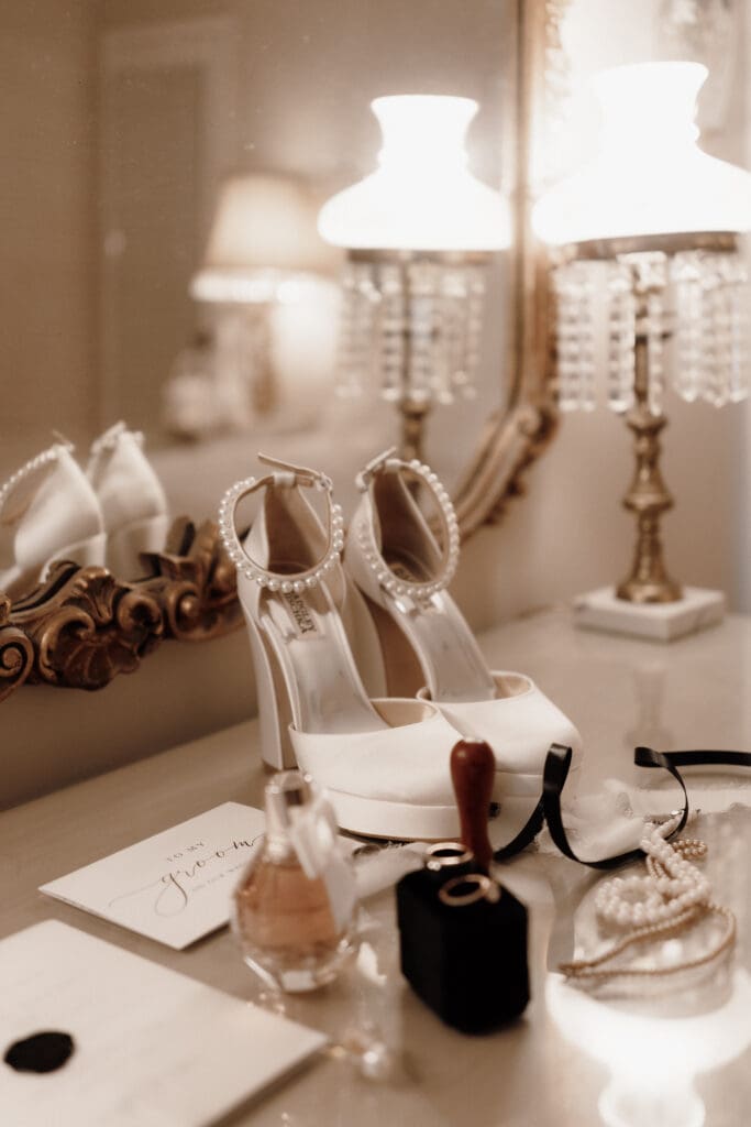Elegant vanity setting featuring white high heels with pearl anklets, surrounded by a vintage lamp, a perfume bottle, a wax seal, a letter, and pearls, reflected in a gilded mirror. Perfect for capturing timeless moments like an Edinburgh wedding photographers dream.