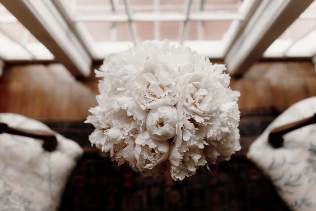 A bouquet of white peonies viewed from above, set in front of large windows, perfect for capturing by a McAllen wedding photographer. The flowers are full and lush, with delicate petals. The interior features a wooden floor and patterned armchairs.