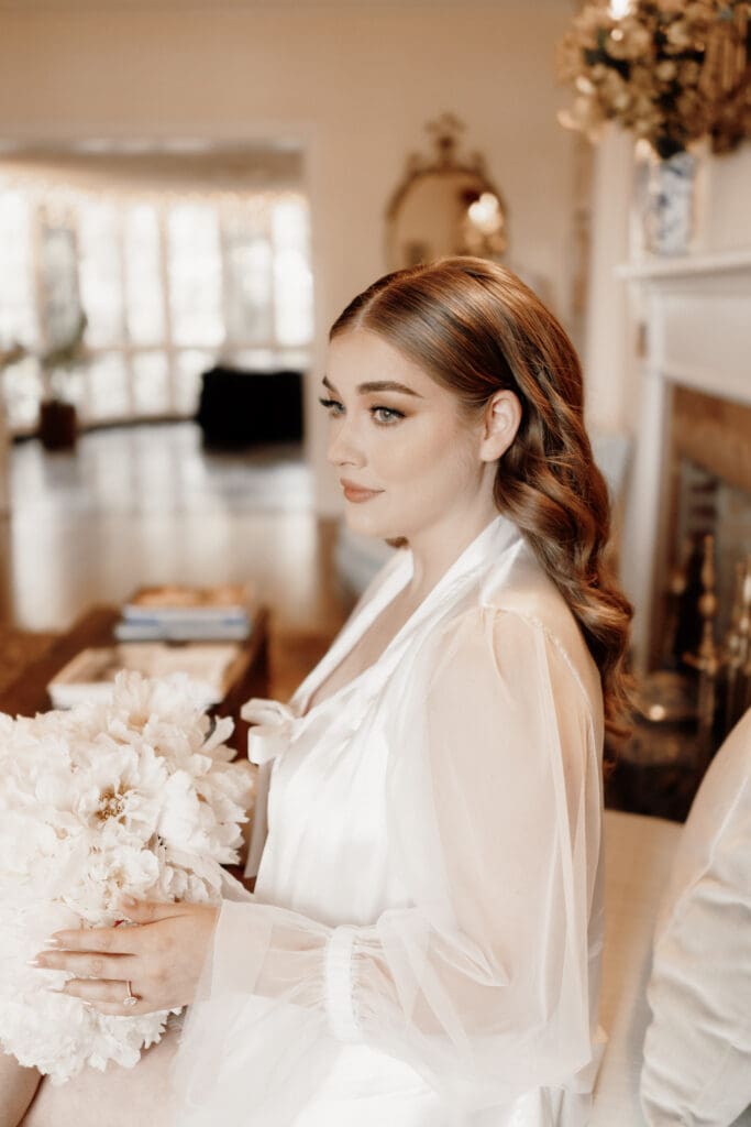 A woman with wavy hair sits indoors holding a bouquet of white flowers. She wears a sheer, long-sleeved top and gazes thoughtfully to the side. Captured by a talented McAllen wedding photographer, the room boasts warm, elegant decor with a mirror and fireplace in the background.