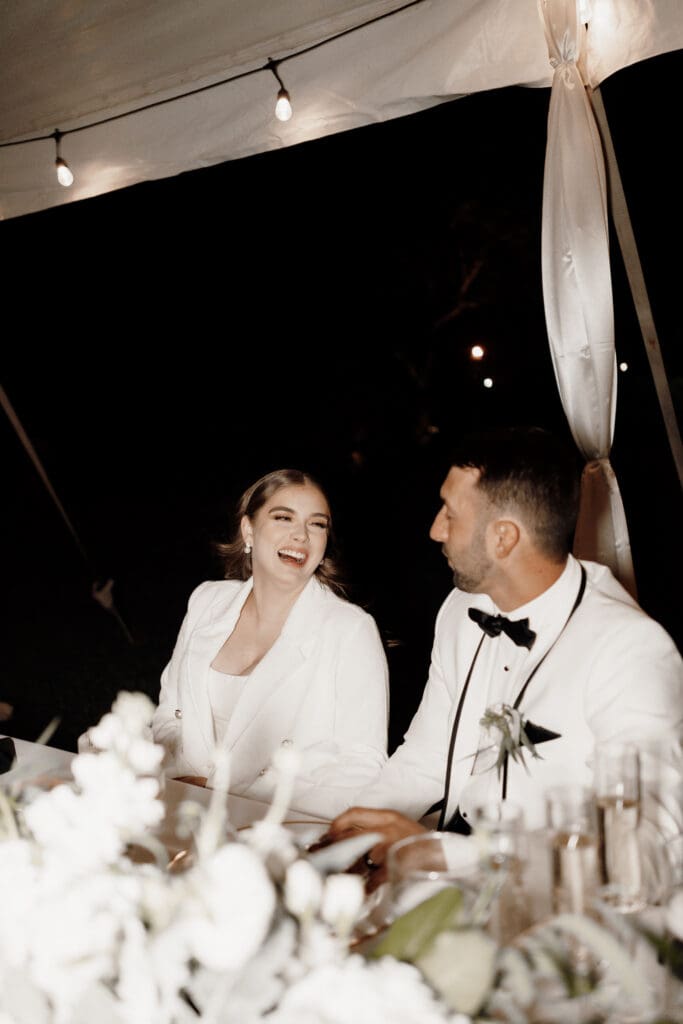 A couple dressed in white sits at a decorated table under a canopy, laughing together. Captured by a talented San Antonio wedding photographer, the enchanting outdoor event sparkles with fairy lights and floral arrangements, creating a magical nighttime setting.