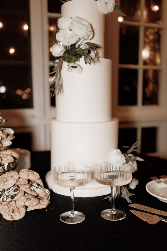 A tall, white wedding cake adorned with floral decorations graces the display. Two ribbed champagne glasses sit elegantly on a black tablecloth before the cake. On the left, a stack of cookies awaits, while to the right rests a gleaming cake server—perfectly captured by a wedding photographer in McAllen.