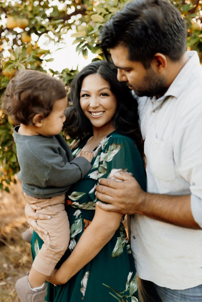 A smiling woman in a green floral dress holds a toddler while a man stands beside her, gently touching her shoulder. Captured by a McAllen wedding photographer, they are outdoors with trees in the background. The family appears happy and relaxed.