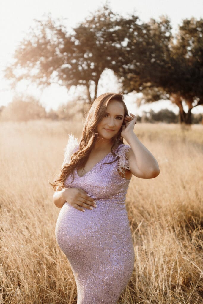 A pregnant woman stands in a sunlit field, her sparkling pink dress shimmering softly. As she gently cradles her belly and brushes her hair, the scene resembles a portrait captured by a skilled Edinburg wedding photographer amid trees and golden grass bathed in warm light.