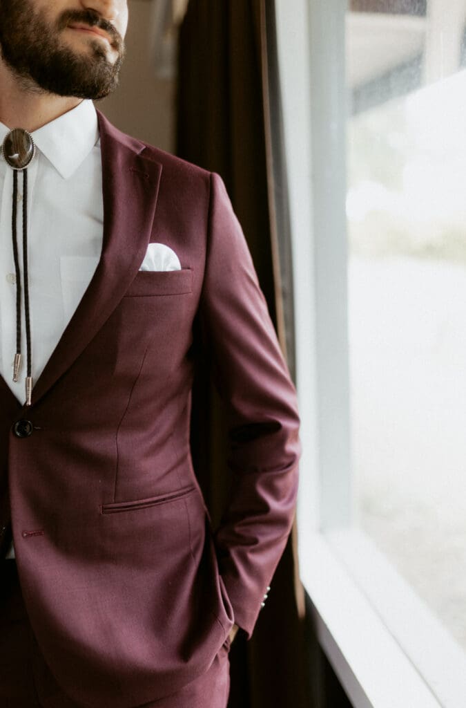 Man in a maroon suit and white shirt with a bolo tie stands near a window, looking outside. His right hand is tucked in his pocket, and a white pocket square is visible. A wedding photographer from McAllen captures the moment, with dark curtains and blurred greenery through the window.